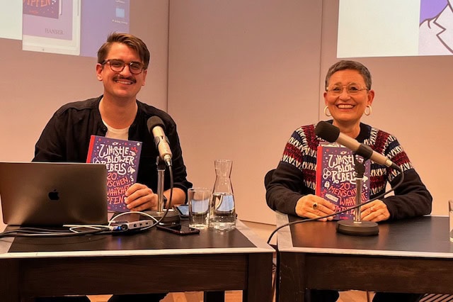 Zwei lächelnde Personen bei einer Lesung, halten ein Buch. Beide sitzen an einem Tisch mit Mikrofon.