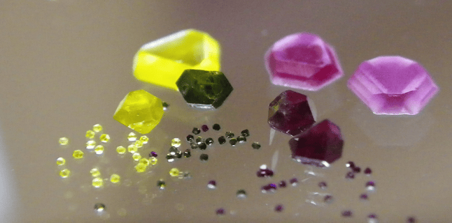 Verschiedene facettierte Edelsteine in Gelb, Grün und Rosa auf einer reflektierenden Oberfläche.