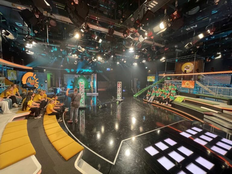 TV-Studio mit Gelb- und Grün-gekleideten Gästen, Sitzreihen, bunten Lichtern, Kameras und Treppen.