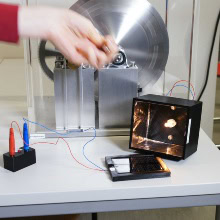 Eine Hand dreht ein Schwungrad; daneben Elektronikbauteile auf weißem Tisch.
