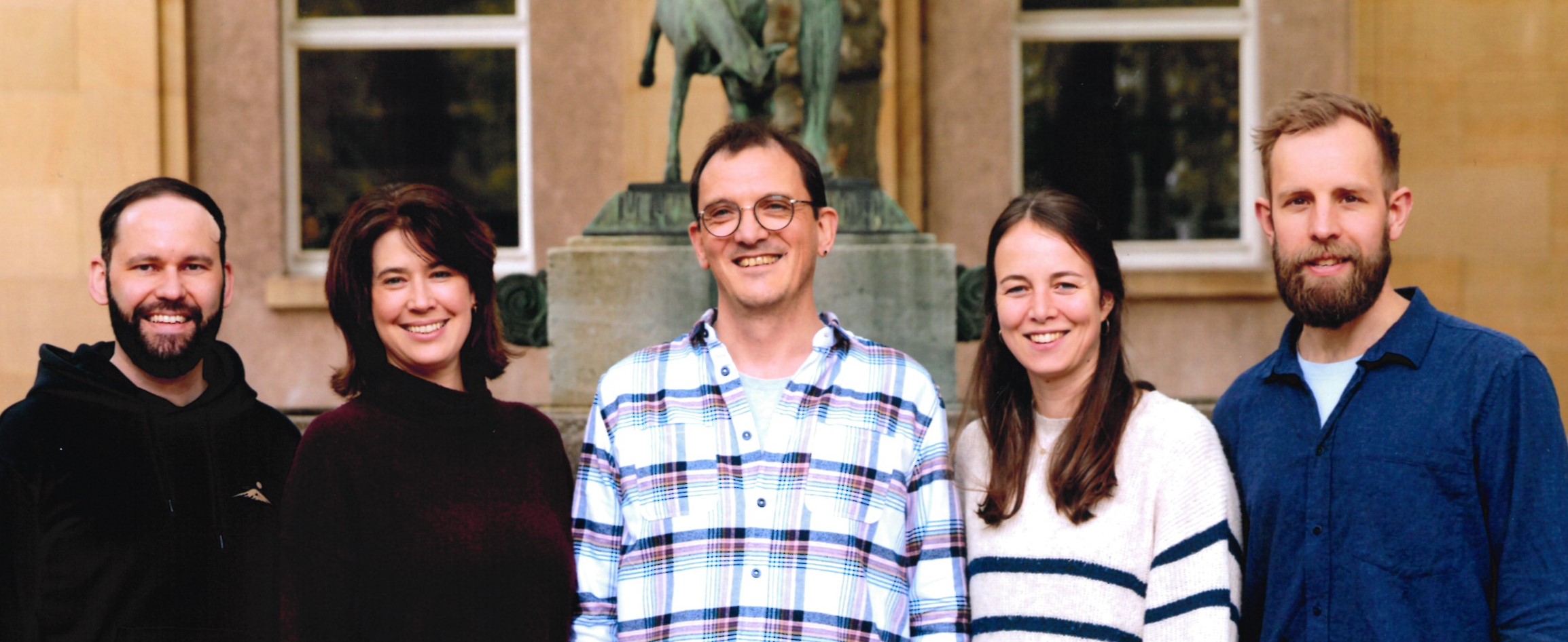 Fünf lächelnde Personen vor einer Statue, zwei Männer mit Bärten, bunt gekleidet.