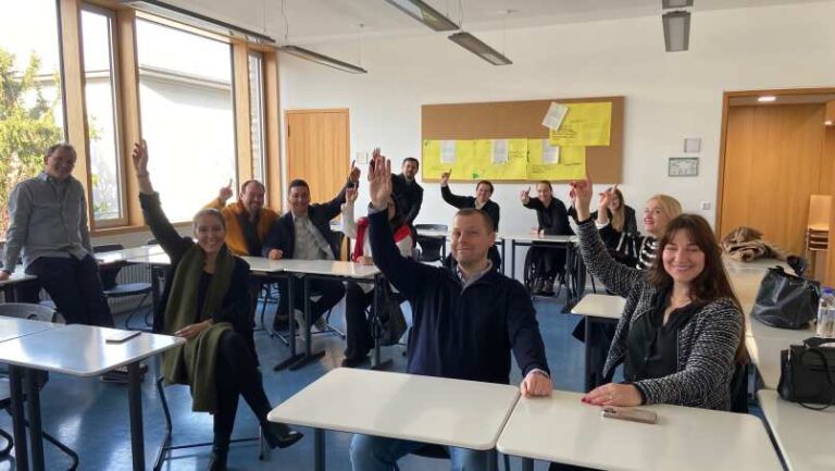 Mehrere Personen sitzen lächelnd in einem Klassenraum und heben die Hand.