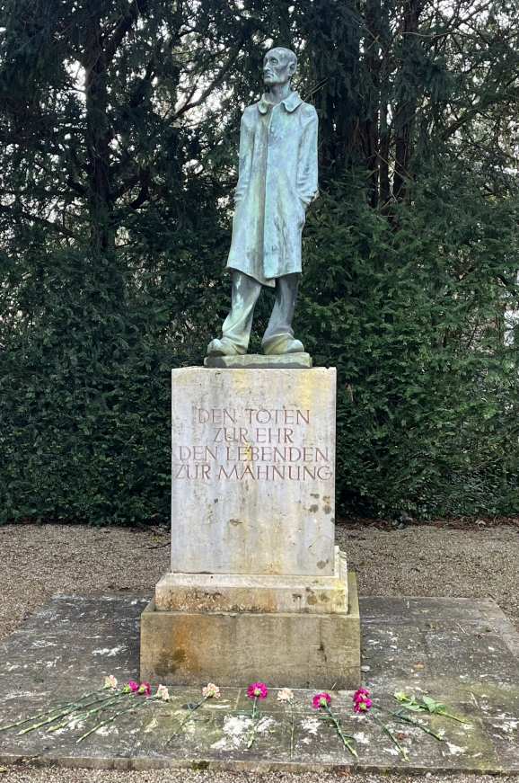 Eine Statue eines Mannes im Mantel, steht ernst, mit Blumen am Sockel auf einer Statue im Park.