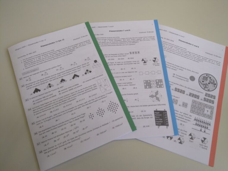Drei farbige Mathematik-Testbögen mit geometrischen Figuren und Aufgaben, auf Tisch liegend.