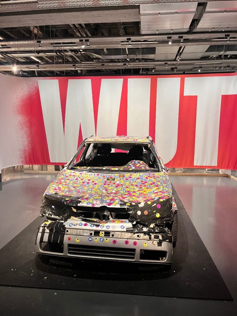 Ein bunt beklebtes Auto vor einer großen, rot-weißen "WUT"-Wand, in einem modernen Raum.