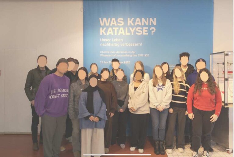 Gruppe von Personen vor blauem Plakat, lächelnd, verschiedene Kleidung und Farben, lebendig.