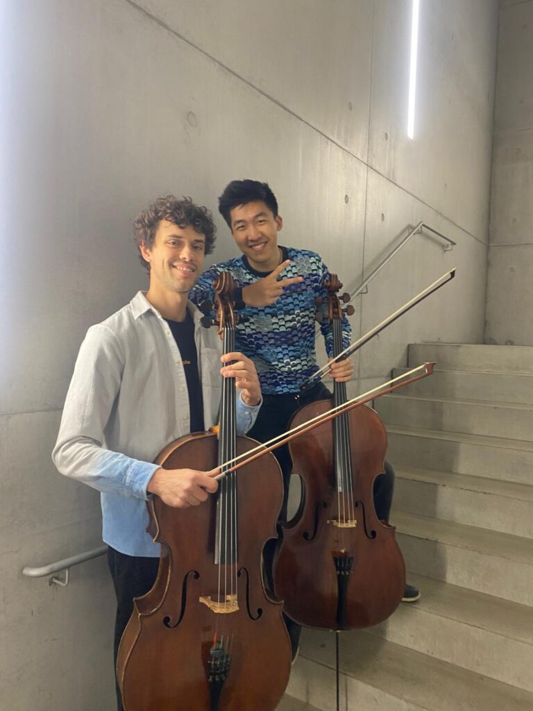 Zwei Männer lächeln mit Cellos auf einer Treppe vor Betonwand.