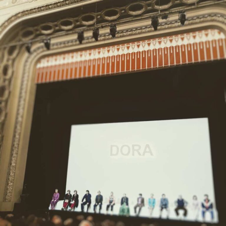 Eine Gruppe von Personen sitzt auf einer Bühne, vor einer Leinwand mit der Aufschrift "DORA".