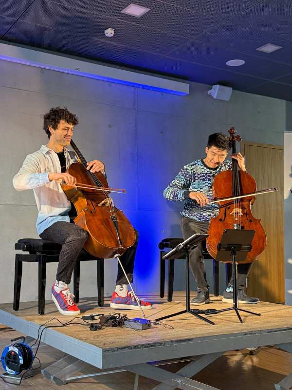 Zwei Musiker spielen lächelnd Cello auf einer Bühne, der Hintergrund ist blau beleuchtet.