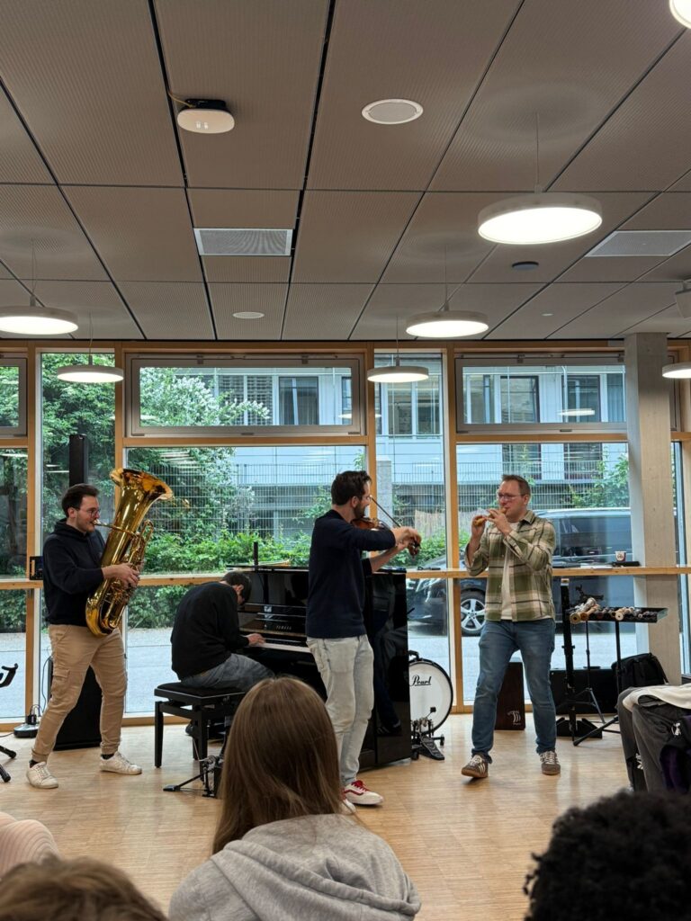 Zwei Männer spielen Tuba und Klavier, einer singt, weiterer klatscht. Raum mit großer Fensterfront.