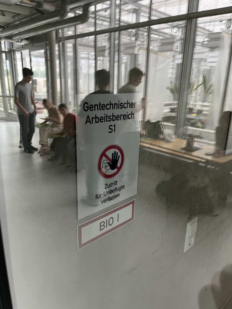 Männer stehen im technischen Bereich durch Glastür mit Warnschild, grauer Boden, Pflanzen sichtbar.