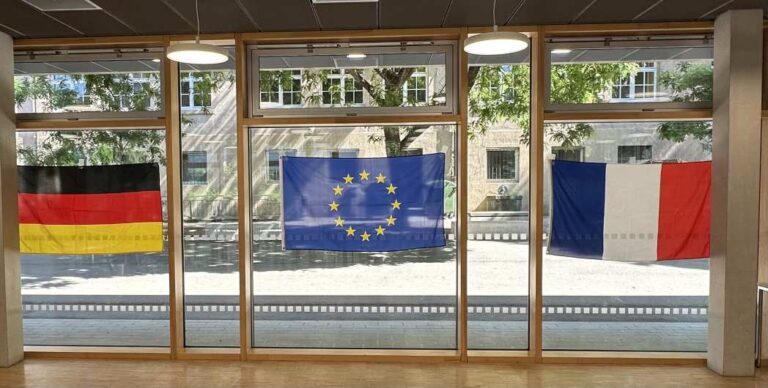Drei Flaggen: Deutschland, EU und Frankreich, hängen vor großen Fenstern in einem modernen Raum.