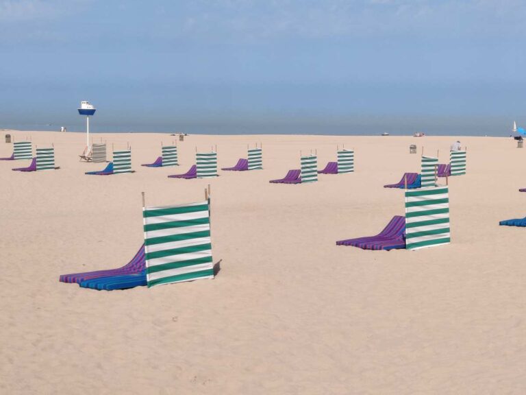 Leerer Strand mit gestreiften Windschutzen und blauen Liegestühlen im Sand.
