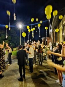 Menschenmenge bei Nacht, gelbe Luftballons steigen in die Luft, freudige Gesichter, festliche Stimmung.