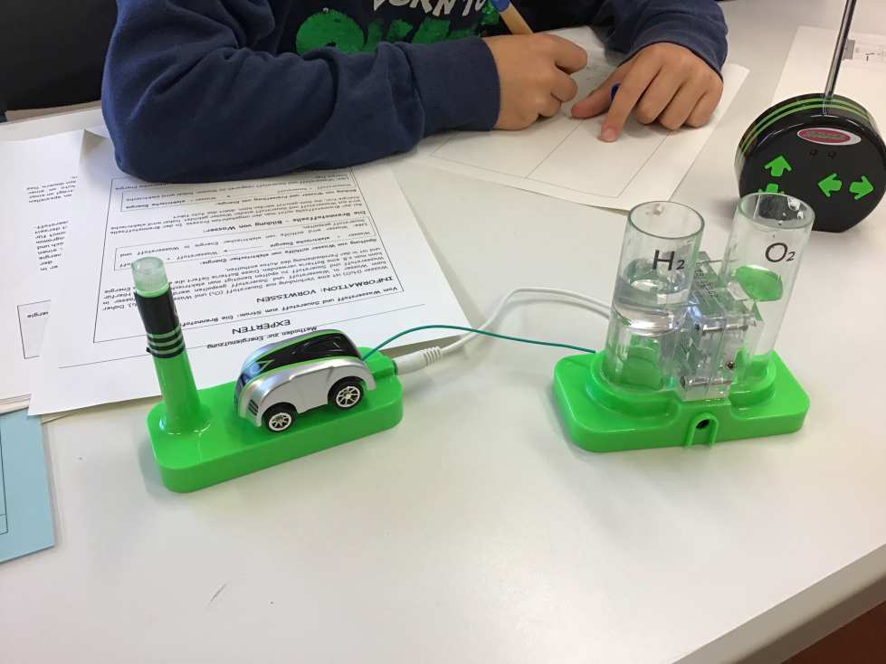 Ein Kind schreibt neben einem Miniaturwasserstoffauto auf einem Tisch mit Experimentaufbau in Grün.