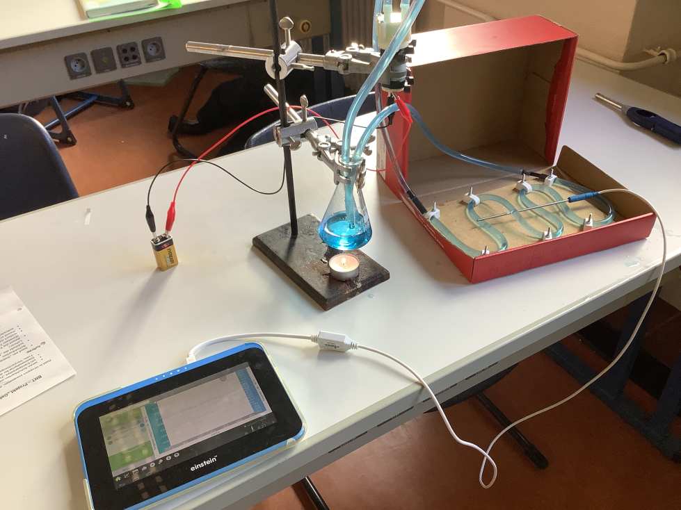 Ein wissenschaftliches Experiment ist auf einem Tisch aufgebaut, mit einem Bunsenbrenner der eine Flüssigkeit in einem Kolben erhitzt, die blau gefärbt ist.