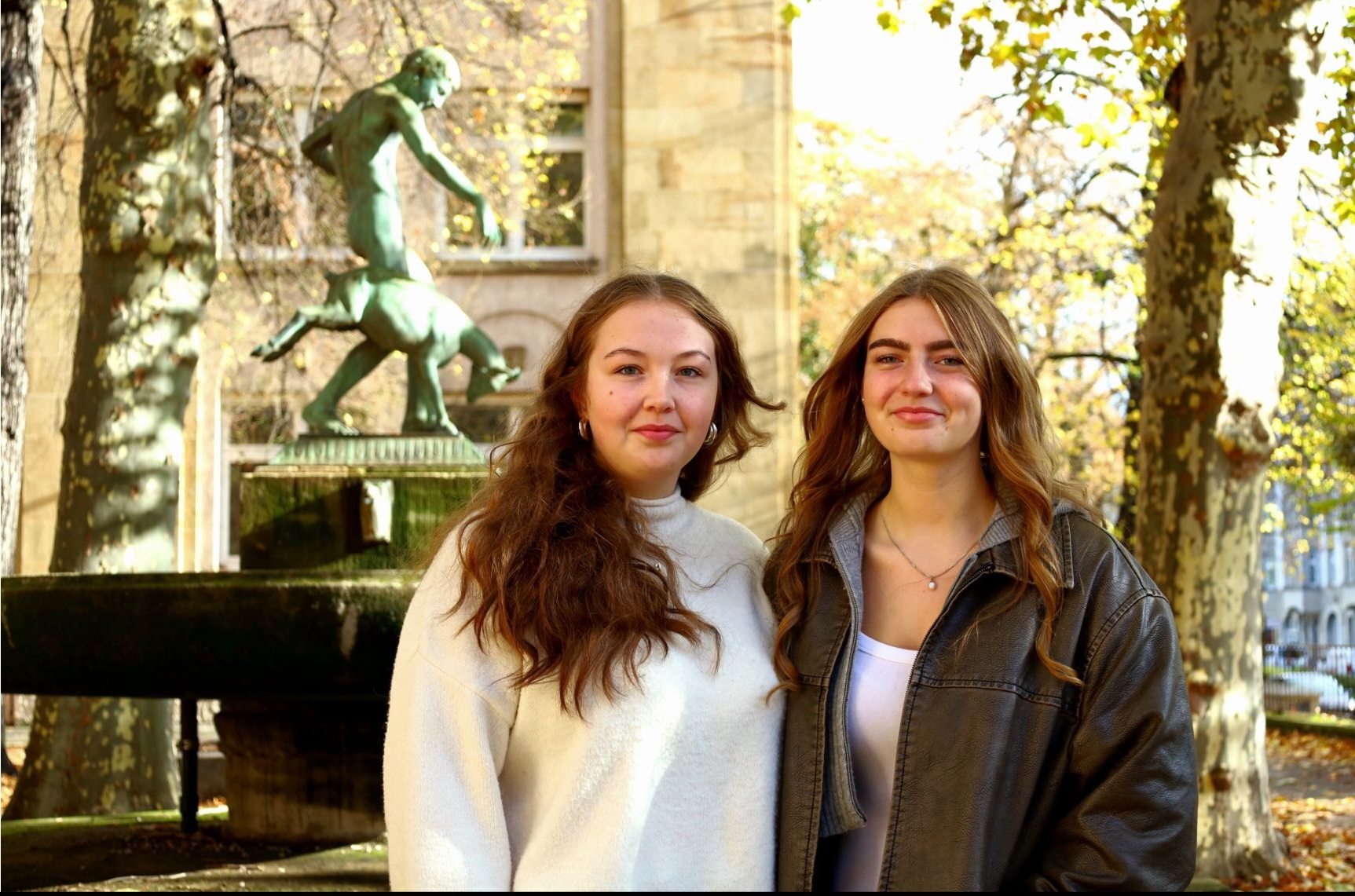 Zwei junge Frauen mit langen braunen Haaren lächeln freundlich im sonnigen Park