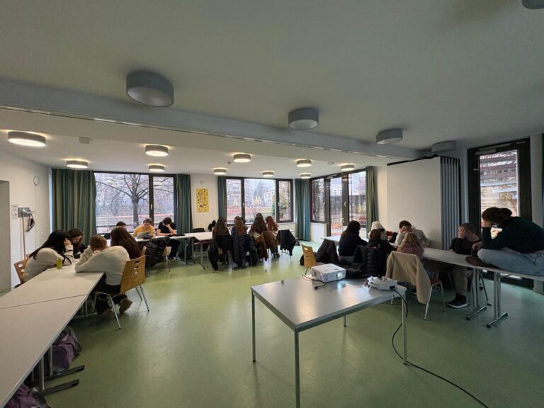 Eine Gruppe Jugendlicher diskutiert an Tischen in einem hellen Klassenraum mit großen Fenstern