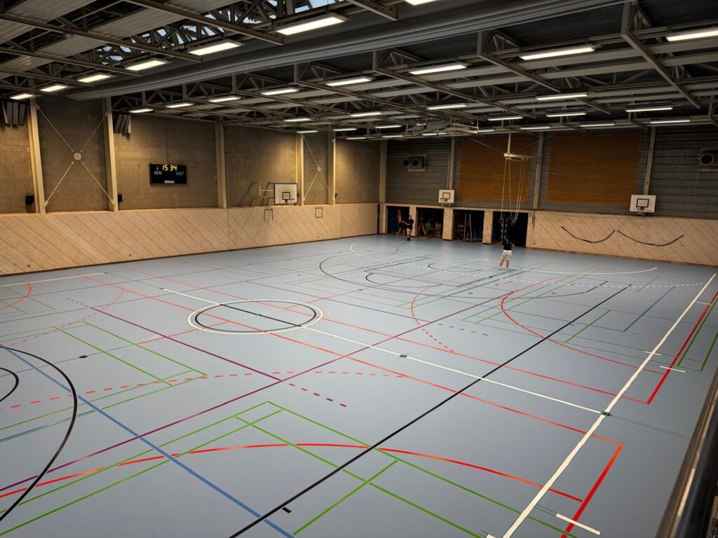 Große leere Sporthalle mit bunten Linien und vereinzelten Personen im Hintergrund