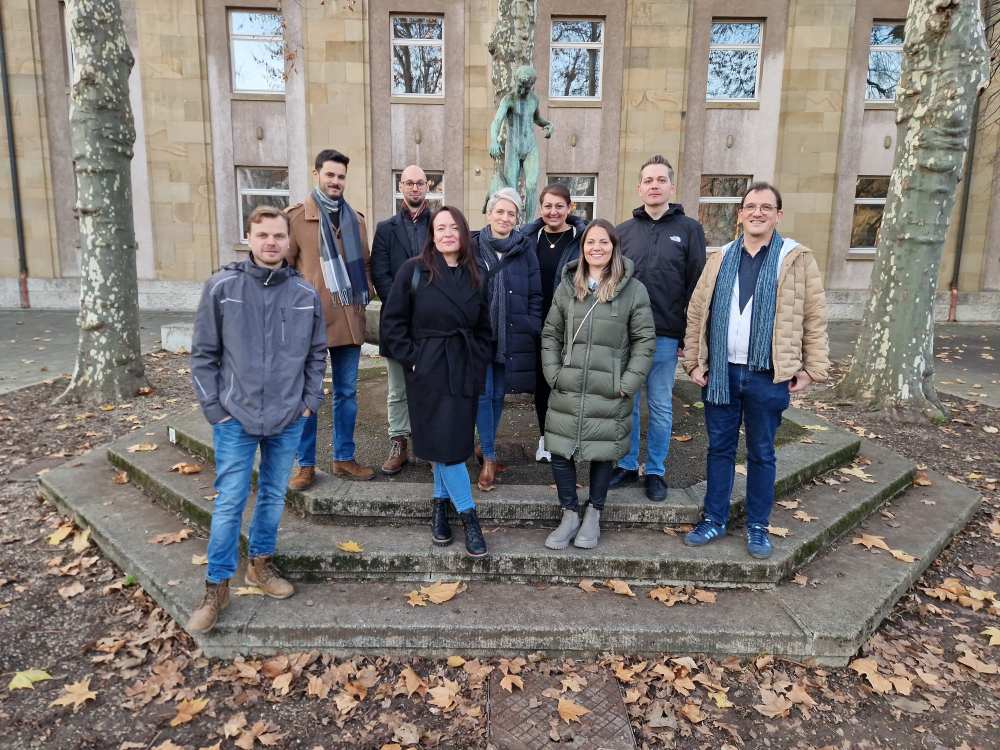 Neun Personen, freundlich lächelnd, stehen auf Steinstufen bei Herbstwetter vor einer Statue