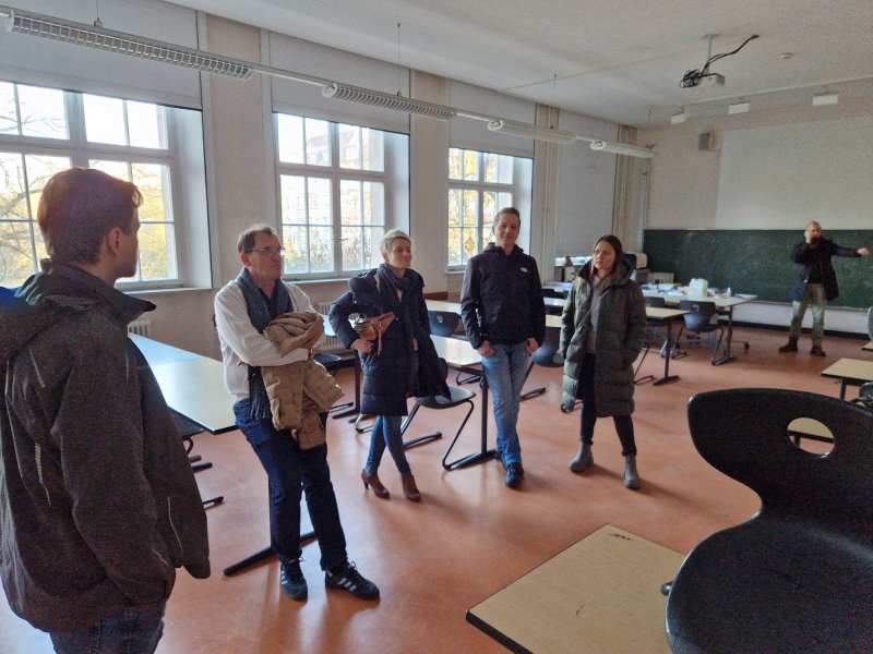 Fünf Erwachsene stehen entspannt plaudernd in einem hellen Klassenraum mit großen Fenstern