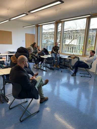 Sieben Personen sitzen aufmerksam in einem modernen Klassenraum mit großen Fenstern