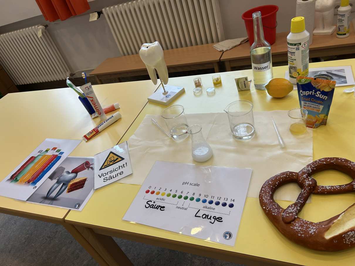 Auf zwei Tischen stehen Zahnpflegeprodukte, Getränke, ein Zahnpmodell und ein Brezel.