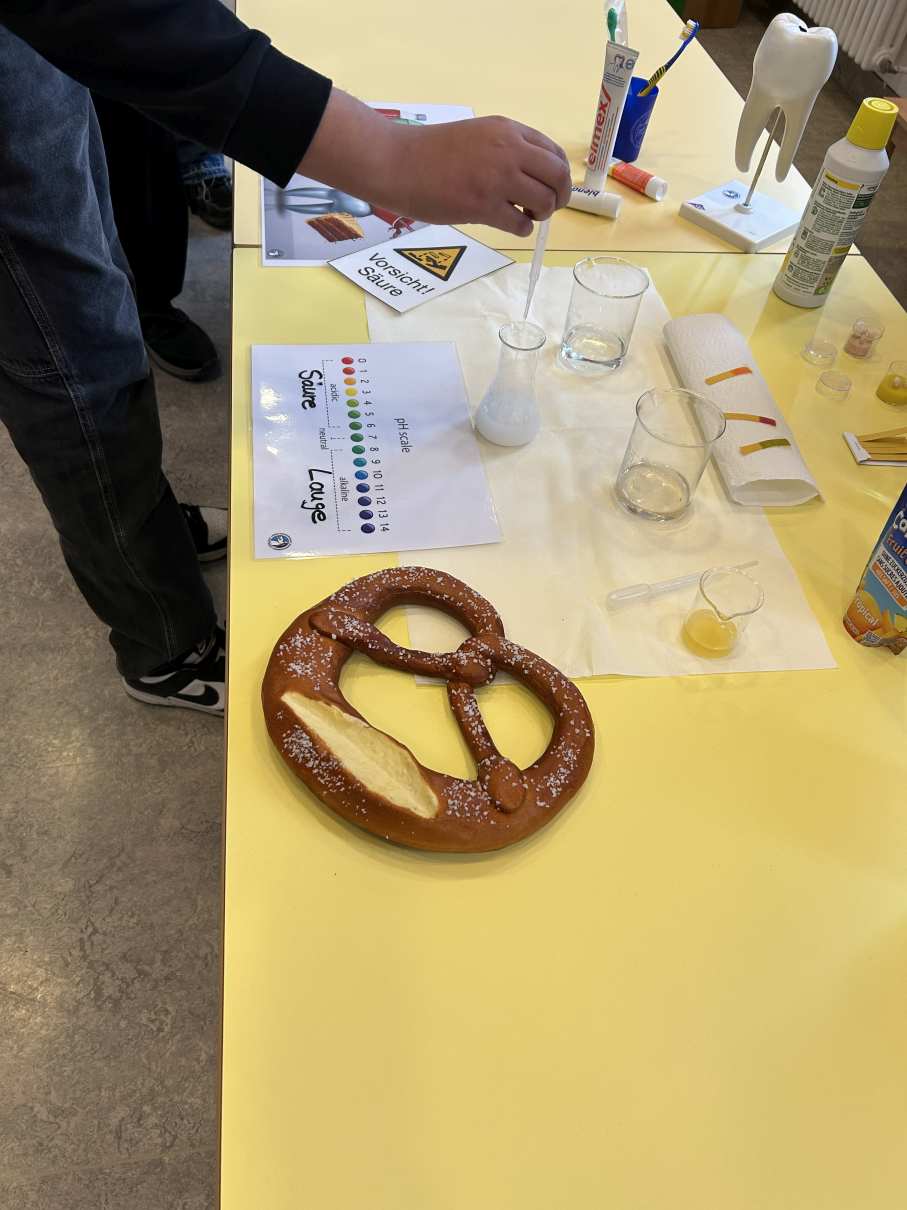 Eine Person mit dunkler Kleidung steht an einem gelben Tisch mit Brezel und Laborutensilien