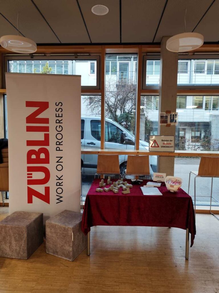 Ein helles Foyer mit einem roten Tisch, Werbebanner von Züblin und kleinen Modellen
