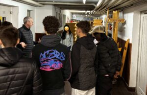 Eine Gruppe junger Menschen hört einem älteren Mann in dunkler Kleidung zu, während sie in einem Flur stehen