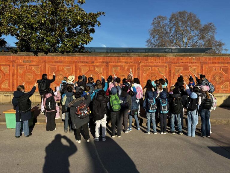 Eine große Gruppe Jugendlicher mit bunten Rucksäcken steht vor einer roten Wand und schaut interessiert