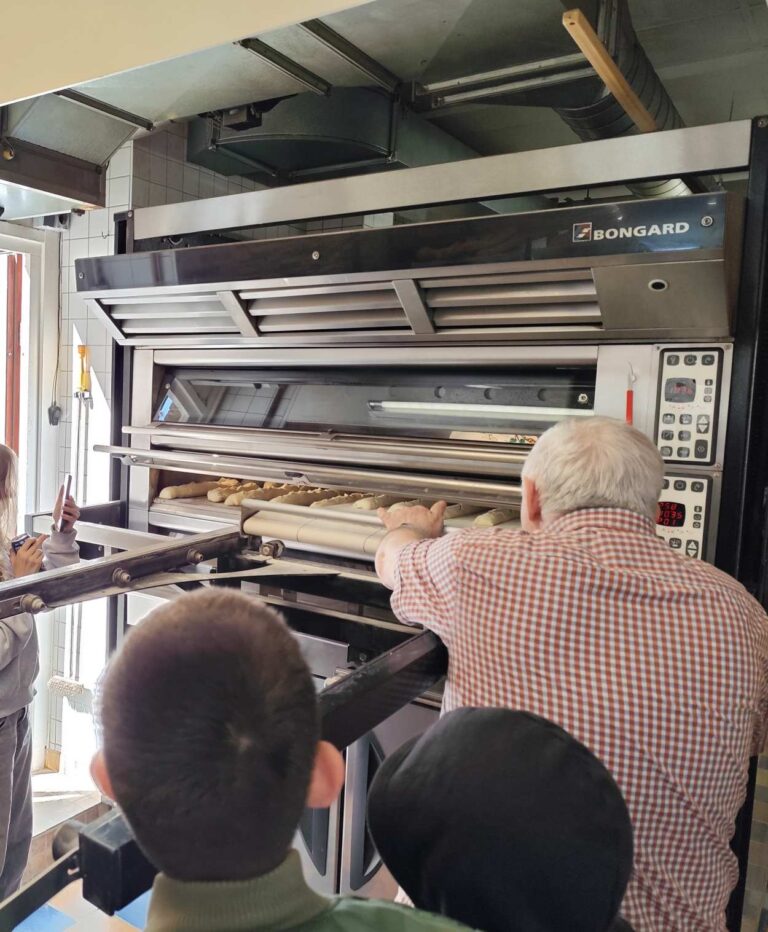 Ein älterer Mann mit weißem Haar schiebt Brot in einen großen silbernen Ofen