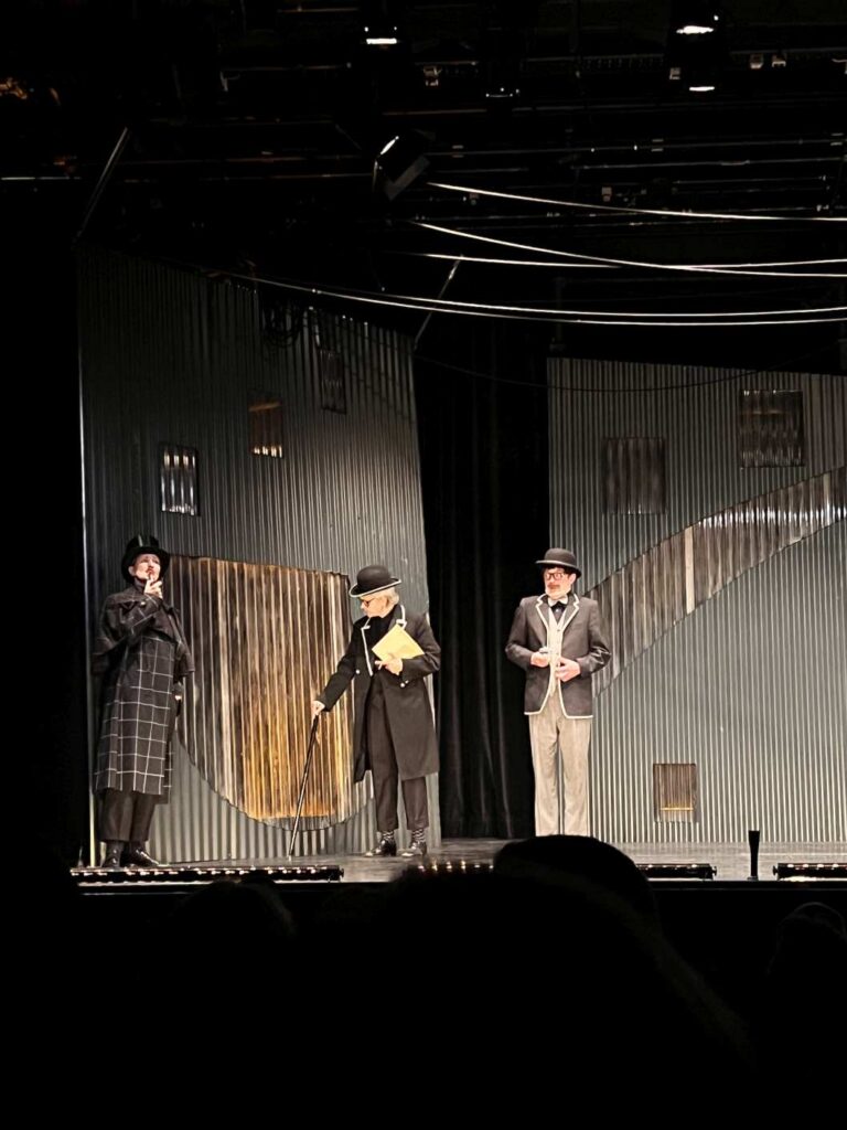 Drei Personen in dunkler Kleidung stehen ernsthaft auf einer Theaterbühne